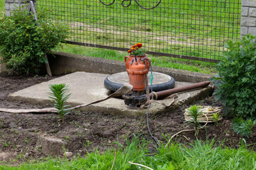 ​A manual water pump stands on a concrete slab, surrounded by soil, plants, and garden tools like a shovel and hose. The pump is adorned with a pot of flowers, creating an atmosphere of simple country