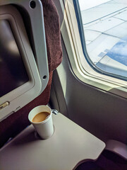 Coffee cup on an airplane tray beside the window, with a clear view of the aircraft wing under bright daylight during flight.