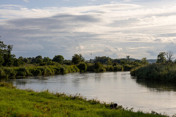 Scenic river landscape with calm water, green banks and cloudy sky in evening light. Peaceful countryside nature view, summer travel and outdoor concept