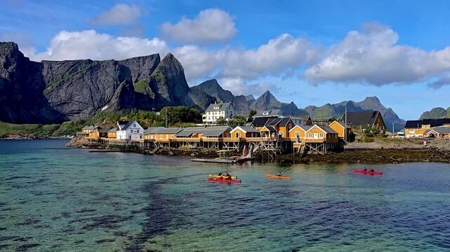 Colorful cabins line the shore as kayakers glide through clear waters in Lofoten, Norway. The majestic mountains rise dramatically, creating a breathtaking landscape perfect for outdoor adventures.
