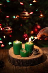 A hand holds a match to light green candles on a wood slice. In the background, there is a decorated Christmas Tree with twinkling lights during the festive New Year holiday.