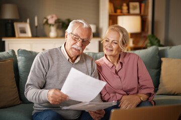 An elderly man wearing glasses reads paperwork while sitting with his wife on a sofa. The wife smiles and looks at the documents with him in a cozy living room setting.