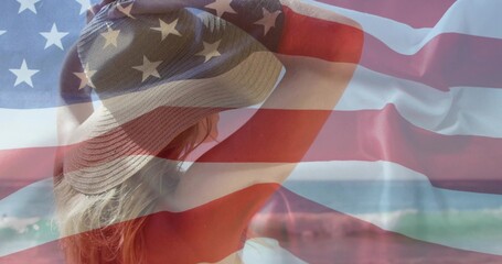 Fototapeta premium Raising bikini-clad female adjusting wide-brimmed sun hat on shore with ocean waves, flag overlay