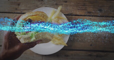 Lifting sesame-seed bun burger with adult hand over wooden tabletop, with ceramic plate of fries