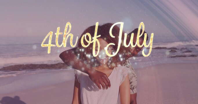 Embracing couple wearing casual tops on sunset beach, showing gold 4th of July text sparkles