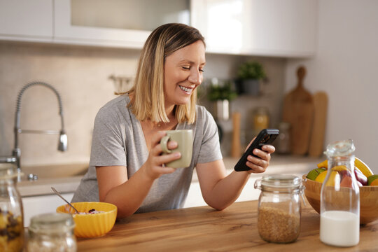 In a warm kitchen, a woman with a joyful expression is holding a mug and looking at her phone. A bowl of cereal and jars of ingredients sit nearby, creating a lively atmosphere.
