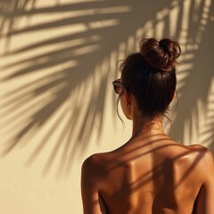 Woman sun-tanned back with shadow of palm leaf. Summer vacation vibes, beach relaxation. Enjoying sunbathing with sunglasses, hair in bun. Minimalist aesthetic, warm light.