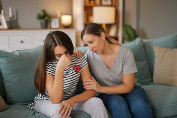 Obraz premium A teenage girl shows distress as she hides her face with her hand, while her mother comforts her with a caring touch on the shoulder in a comfortable living room.
