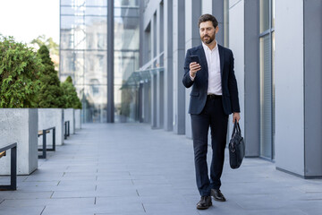Confident businessman in a suit walking with a briefcase and smartphone near a modern office building in the financial district, embodying urban lifestyle and business communication
