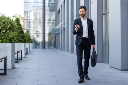 Confident businessman in a suit walking with a briefcase and smartphone near a modern office building in the financial district, embodying urban lifestyle and business communication
