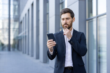 Businessman in a suit is standing near an office building, holding his smartphone and looking thoughtfully at the screen while stroking his beard