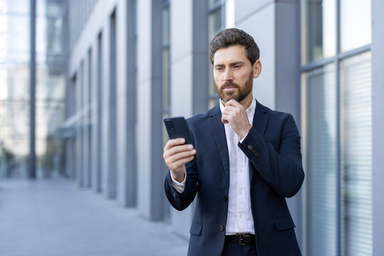 Businessman in a suit is standing near an office building, holding his smartphone and looking thoughtfully at the screen while stroking his beard