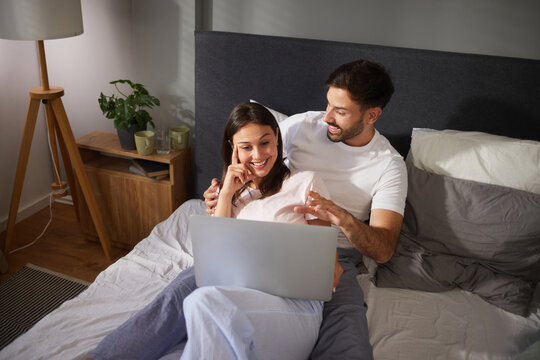 A couple enjoys planning their vacation together, sitting on a bed while using a laptop. They appear engaged and excited about their travel options, surrounded by a warm atmosphere.
