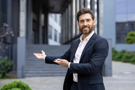 Happy businessman in a suit, smiling and welcoming, gesturing with his hands in front of a modern office building, exuding confidence and professionalism in an urban setting