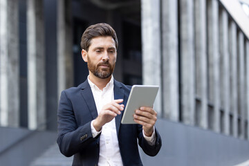 Bearded businessman standing confidently in front of an office building, using a digital tablet to work remotely, showcasing modern technology and efficient communication in an urban setting