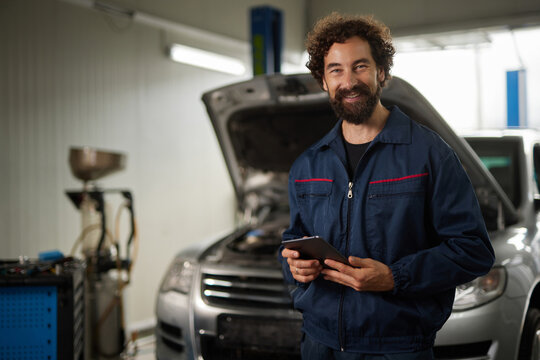 A car mechanic smiles while holding a tablet inside a repair garage. The engine of a vehicle is visible under the raised hood, showcasing ongoing maintenance work.