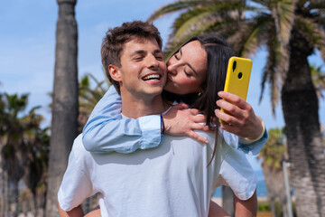Happy young couple outdoors with palm trees, woman hugging and kissing man while holding yellow smartphone for selfie, lifestyle, romance, leisure, modern love, online connection, fun summer activity