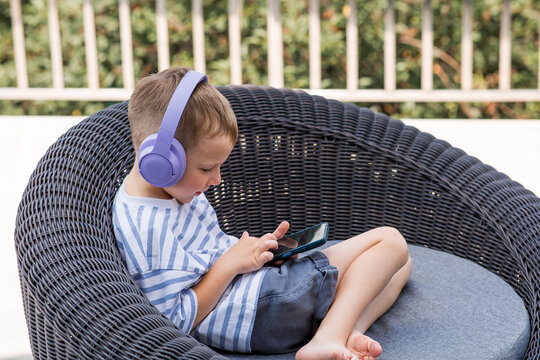 A 6-year-old boy is sitting on the couch with a smartphone in his hands and purple headphones on. The child is playing and listening to music. Children and gadgets.