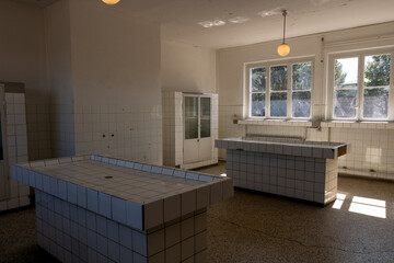 Empty tiled room with large counter tables, windows letting in daylight, and minimal furnishing, creating a stark and utilitarian interior atmosphere.