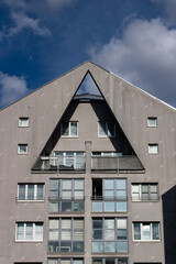 Modern residential building with triangular roof design, sharp geometric lines, large windows, and balconies under a bright blue sky with clouds.