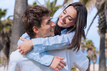 Happy young couple outdoors with palm trees, woman hugging man from behind, lifestyle, romance, leisure, modern love, summer fun, confidence, affection, positive emotions, stylish casual people © Raquel