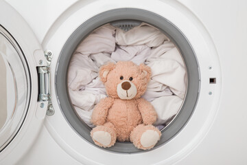 Soft teddy bear sitting in washing machine. Baby white clothes washing. Closeup. Front view.