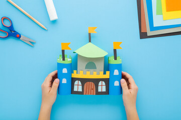 Little child boy hands holding colorful castle with towers from paper. Scissors, glue stick, pencil and application paper on light blue table background. Pastel color. Closeup. Top down view.
