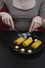 Woman enjoying stuffed crepe rolls served on a black plate with decorative sauce dots.