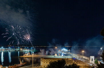 Fireworks light up the night sky over a bustling waterfront, reflecting on the water as crowds gather to celebrate in a vibrant urban coastal scene.
