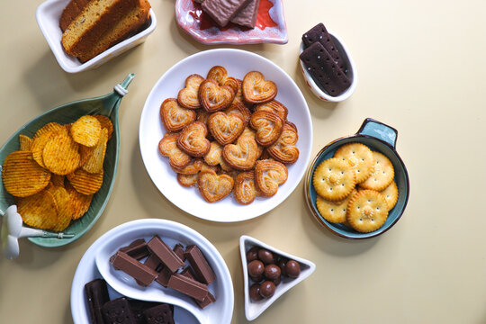 Assorted party snacks on table featuring chocolate cream sandwich biscuits, sugar coated heart cookies, wafer chocolates, chocolate coated caramel balls, salted crackers, and crispy potato chips.