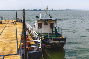 Obraz premium Rustic small working boat docked at a wooden pier on a calm river or lake. Old vessel with simple cabin and ropes, representing river transport and fishing lifestyle.