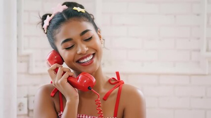 Joyful young woman with dark hair wearing colorful hair accessories, smiling while talking on a vintage red telephone against a bright background - Powered by Adobe