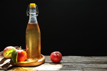 Delicious cider in glass bottle and apples on wooden table against dark background. Space for text