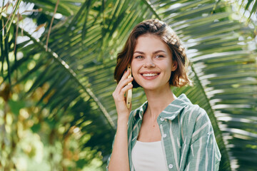 woman, portrait, smile, outdoors, phone call, casual clothes, green foliage, sunny day, cheerful mood, friendly gesture, natural light, candid moment, relaxed vibe, warm surroundings, bright