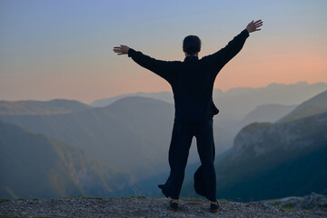Woman practicing Tai Chi in traditional black outfit on mountain at sunrise, arms outstretched in meditation pose focusing on balance, energy, and mindfulness in nature.