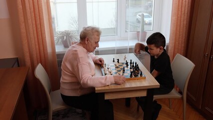 Grandmother playing chess with grandson at the table by the window. A good way to spend time - Powered by Adobe