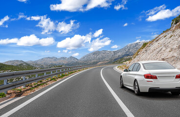 A white car drives along a curving mountain highway under a bright blue sky with scattered clouds,...