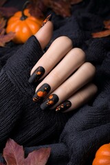 Una mano femminile con unghie decorate con motivi di zucca color arancione su uno sfondo nero lucido, circondato da zucche e foglie autunnali per festeggiare  Halloween.