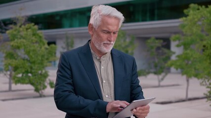 Confident senior businessman with gray hair using tablet in city office area. He is wearing smart casual outfit and appears focused in urban business district - Powered by Adobe