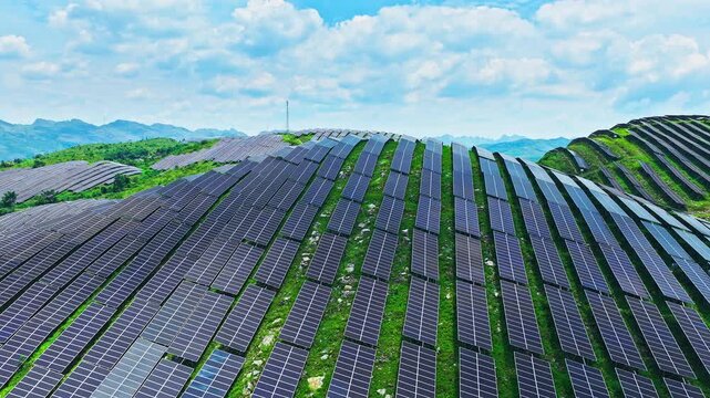 Aerial shot of long rows of solar panels curving over a green hillside on a beautiful sunny day with a blue sky.