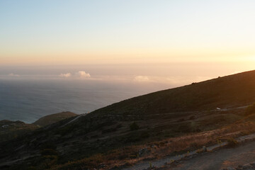 Obraz premium A scenic view of a hillside overlooking the ocean at sunset. The sky is a gradient of orange and blue, with distant hills visible across the water.
