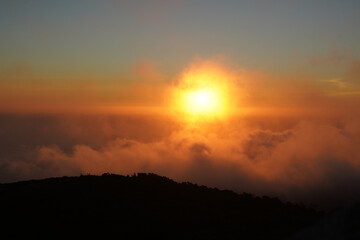A vibrant sunset glows over a landscape of clouds and a silhouetted mountain. The sun's rays pierce through the clouds, creating a dramatic and colorful scene.