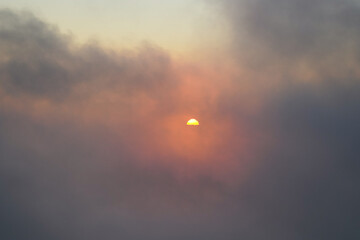 A hazy sunset emerges from behind dark clouds. The sun is a small, bright orb, surrounded by orange and pink hues, creating a dramatic and moody sky.