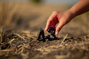 A woman's hand places black candles in the shape of the number forty-three into the soil of a cultivated agricultural field
