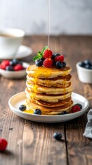 Stack of golden pancakes drizzled with honey, topped with fresh raspberries and blueberries, on a wooden table