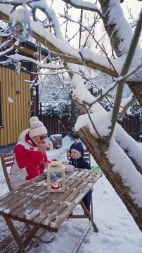 Grandmother and child enjoying tea outdoors in Finland winter, wearing warm seasonal clothing, cozy Nordic lifestyle in snowy nature.