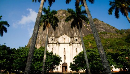 Obraz premium A serene, light-filled view of a historical church nestled beside a massive rock face, shaded by tall palm trees.
