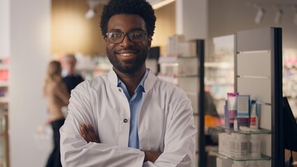 African American ethnic man portrait health care worker doctor pharmacist smiling guy in pharmacy drugstore shelves with medicine male physician seller in medical coat healthy insurance crossed hands