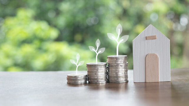 Stacked coins with plant sprouts and a wooden house model, symbolizing sustainable financial growth, real estate investment, savings, green finance, and an eco-friendly property concept.