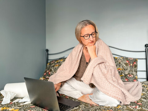 Middle-aged woman sick at home with cold and flu symptoms. Sitting on bed wrapped in a soft blanket, surrounded by tissues, pills and thermometer. Working remotely on laptop.